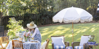 Woman in a straw hat arranging flowers on a blue-and-white table set for outdoor spring dining, with wicker chairs, striped umbrella, and garden seating on a sunny lawn.
