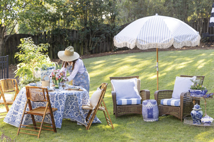 Woman in a straw hat arranging flowers on a blue-and-white table set for outdoor spring dining, with wicker chairs, striped umbrella, and garden seating on a sunny lawn.