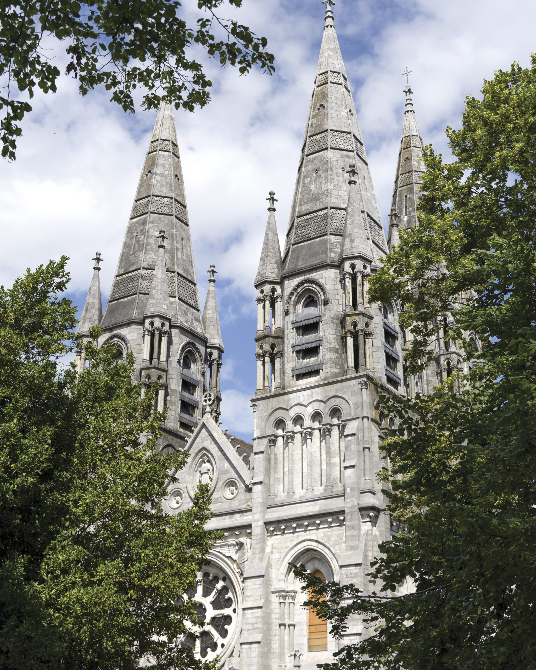 Alt text: Stone Gothic Revival church with three tall spired towers rising above leafy green trees, featuring arched windows, decorative stonework, and a rose window beneath a partly cloudy blue sky.