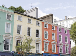 Colorful pastel row houses in Cobh, Ireland—painted mint green, pale yellow, coral orange, and pink—line a sunny street with café storefronts below and white-trimmed windows above.