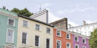 Colorful pastel row houses in Cobh, Ireland—painted mint green, pale yellow, coral orange, and pink—line a sunny street with café storefronts below and white-trimmed windows above.