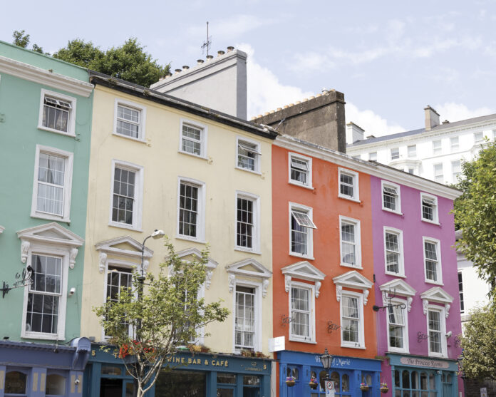 Colorful pastel row houses in Cobh, Ireland—painted mint green, pale yellow, coral orange, and pink—line a sunny street with café storefronts below and white-trimmed windows above.