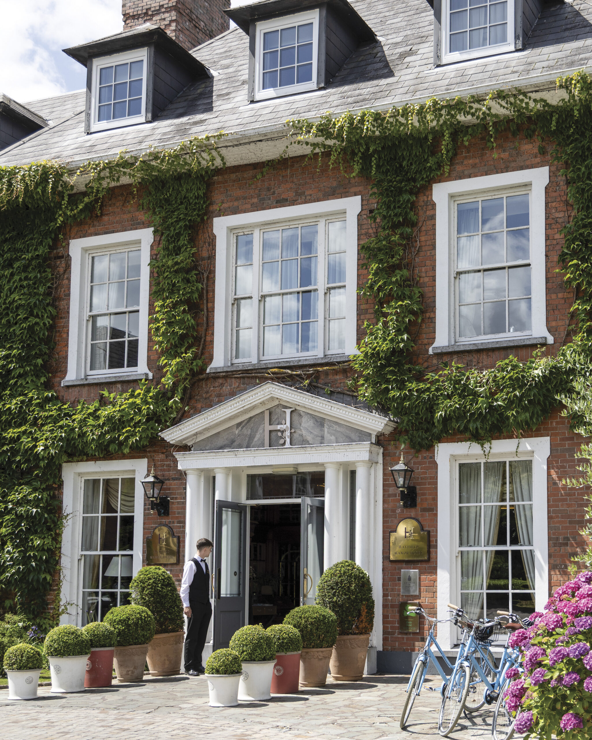 Ivy climbs the red-brick façade of Hayfield Manor in Cork, Ireland, where a staff member stands at the columned entrance beside potted topiaries and bicycles resting near blooming hydrangeas.