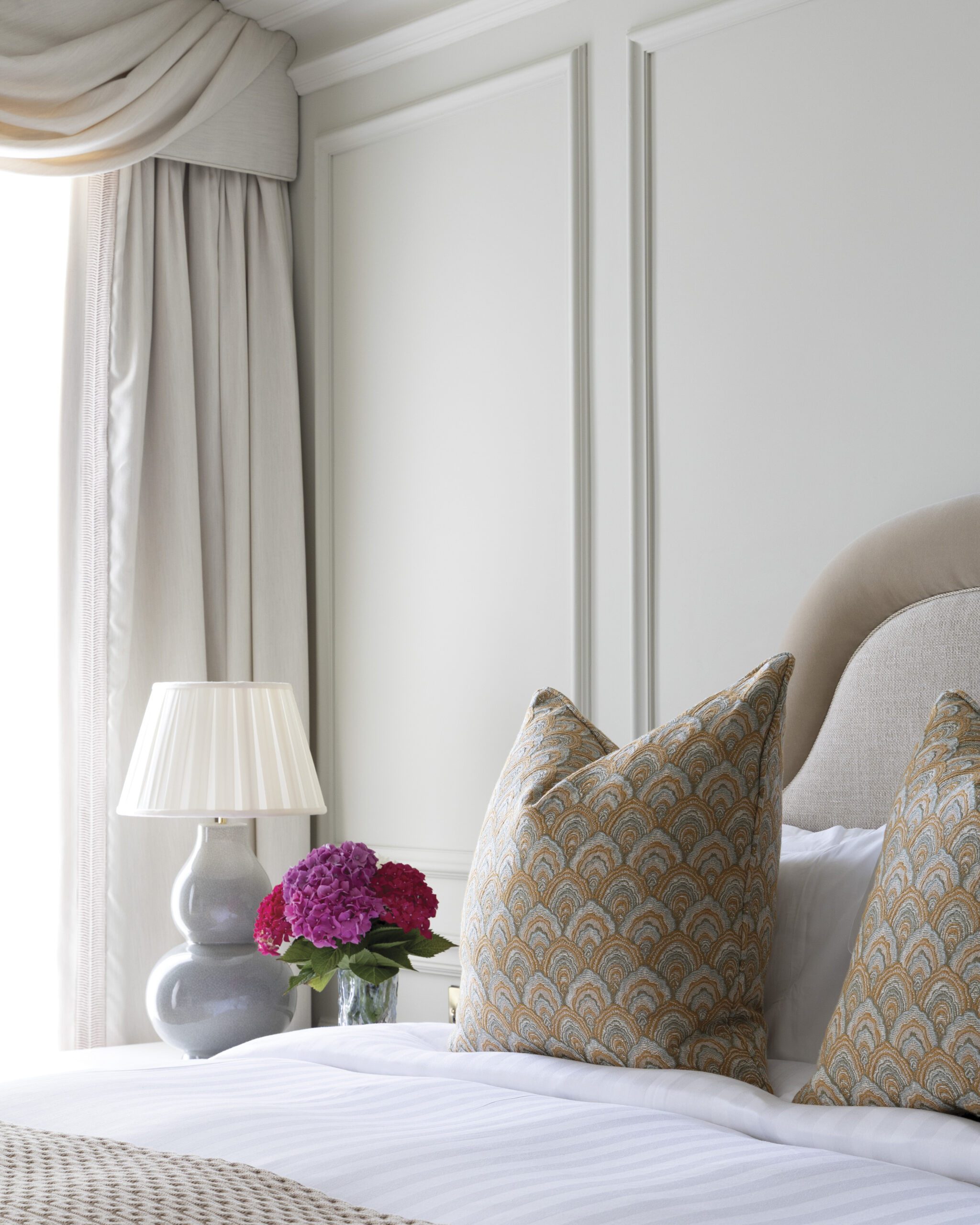Elegant guest room at Hayfield Manor in Cork featuring a cream upholstered headboard, patterned accent pillows, soft draped curtains, and a bedside table with a ceramic lamp and pink hydrangeas.