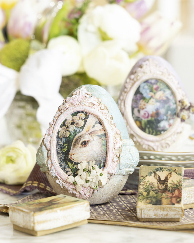 Two decorative Easter eggs featuring vintage-style rabbit illustrations in ornate frames sit on a tray, surrounded by soft florals and small keepsake boxes in a pastel spring arrangement.