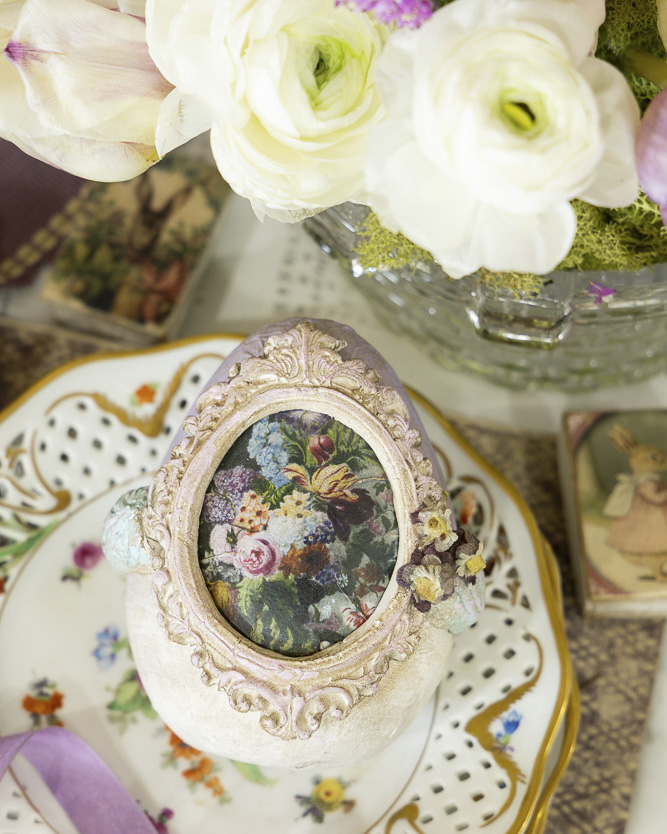 A decorative Easter egg adorned with an ornate oval floral medallion sits atop a pierced porcelain plate with delicate hand-painted flowers, surrounded by fresh blooms and vintage-style keepsakes.