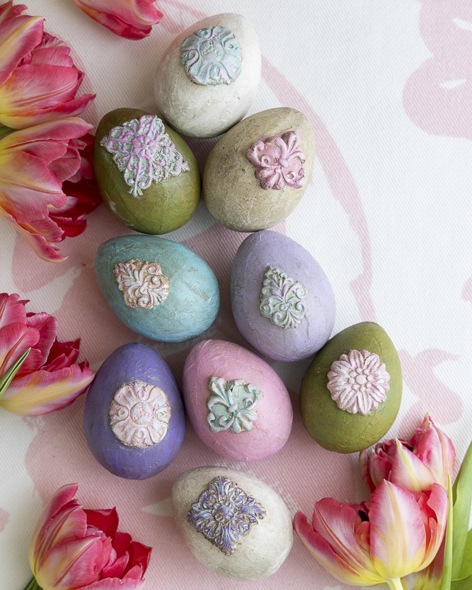 A collection of pastel-painted Easter eggs adorned with raised ornamental designs, arranged on a soft pink patterned surface and framed by vibrant tulips.