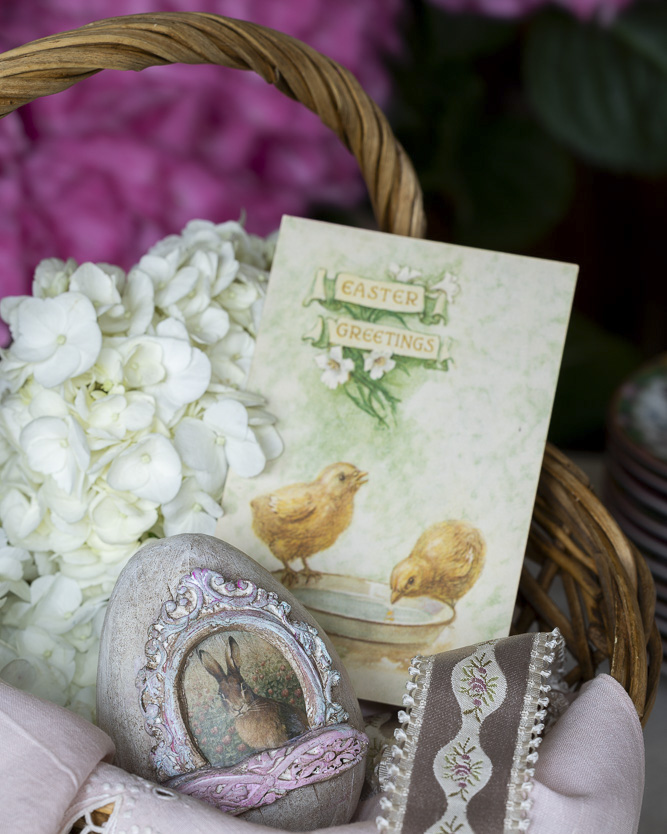 A woven basket filled with white hydrangeas, a vintage-style Easter card with chicks, and a decorative egg featuring a framed rabbit illustration, arranged with soft pink ribbon and embroidered trim.