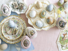 An overhead view of an elegant Easter tablescape featuring pastel eggs arranged in decorative nests on ornate china plates, surrounded by vintage postcards and embellished eggs with floral and rabbit motifs.