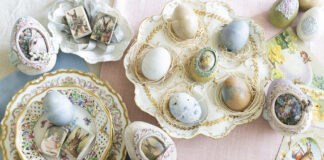 An overhead view of an elegant Easter tablescape featuring pastel eggs arranged in decorative nests on ornate china plates, surrounded by vintage postcards and embellished eggs with floral and rabbit motifs.