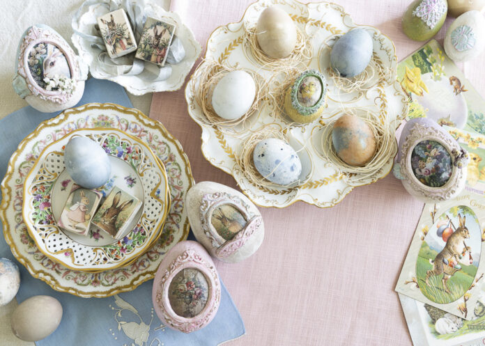 An overhead view of an elegant Easter tablescape featuring pastel eggs arranged in decorative nests on ornate china plates, surrounded by vintage postcards and embellished eggs with floral and rabbit motifs.
