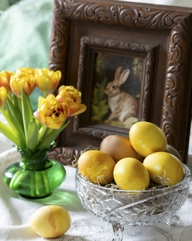 A spring vignette with yellow-dyed eggs nestled in a cut-glass bowl, a green vase of yellow tulips, and a framed rabbit illustration, arranged on a lace-covered surface.