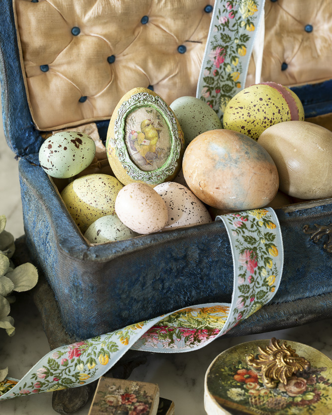 A velvet-lined antique box filled with pastel and speckled Easter eggs, including one with a vintage chick illustration, accented by a floral embroidered ribbon.