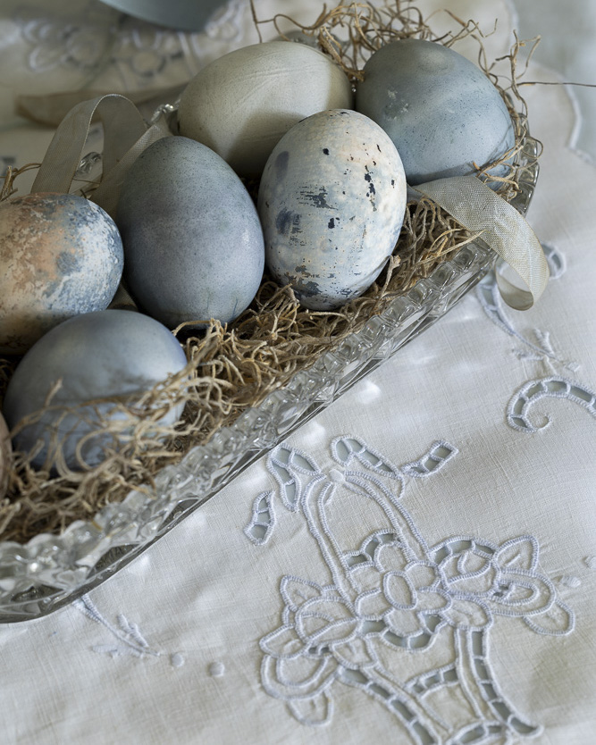 Naturally dyed eggs in muted blue and gray tones rest in a cut-glass dish lined with dried moss and ribbon, set atop an embroidered white linen cloth.
