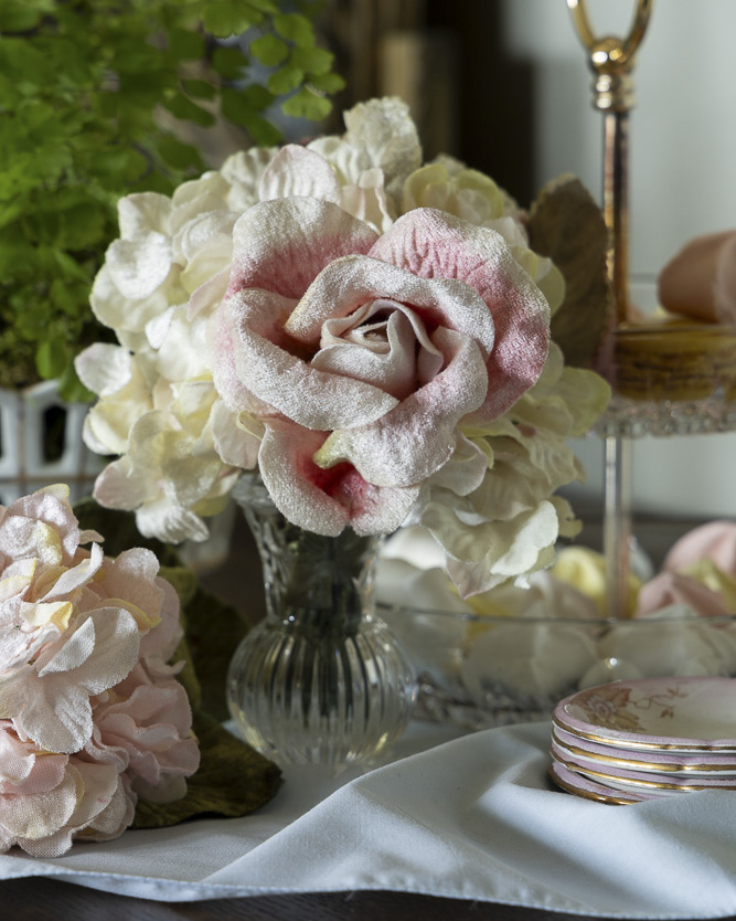 A soft arrangement of blush and ivory flowers in a glass vase sits beside delicate pink-rimmed plates and a tiered stand, creating an elegant, romantic tabletop vignette.