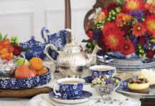 A richly styled tea table with blue-and-white china teacups, a silver teapot, fresh fruit, and petite desserts, arranged alongside a vibrant bouquet of red, orange, and purple flowers.