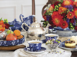 A richly styled tea table with blue-and-white china teacups, a silver teapot, fresh fruit, and petite desserts, arranged alongside a vibrant bouquet of red, orange, and purple flowers.