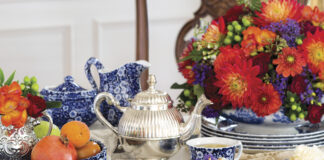 A richly styled tea table with blue-and-white china teacups, a silver teapot, fresh fruit, and petite desserts, arranged alongside a vibrant bouquet of red, orange, and purple flowers.