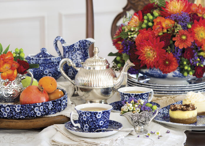 A richly styled tea table with blue-and-white china teacups, a silver teapot, fresh fruit, and petite desserts, arranged alongside a vibrant bouquet of red, orange, and purple flowers.