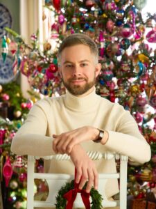 Smiling man with a beard in a cream sweater leaning on a white chair, with colorful Christmas ornaments behind him.