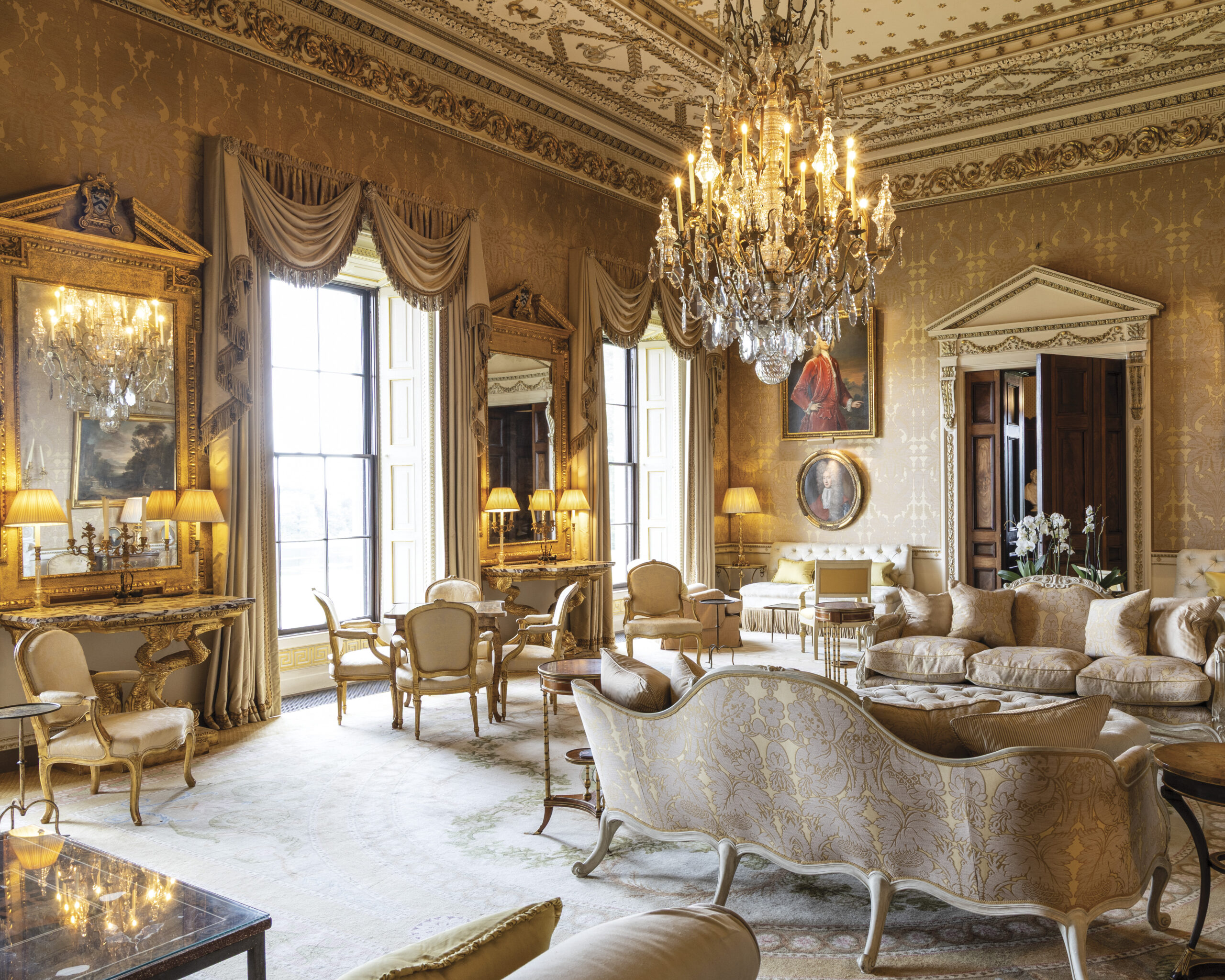 An opulent, gold-toned salon features ornate plasterwork ceilings, crystal chandeliers, and tall windows draped in silk, with gilded mirrors, antique seating, and elegant furnishings arranged in a refined, classical interior.