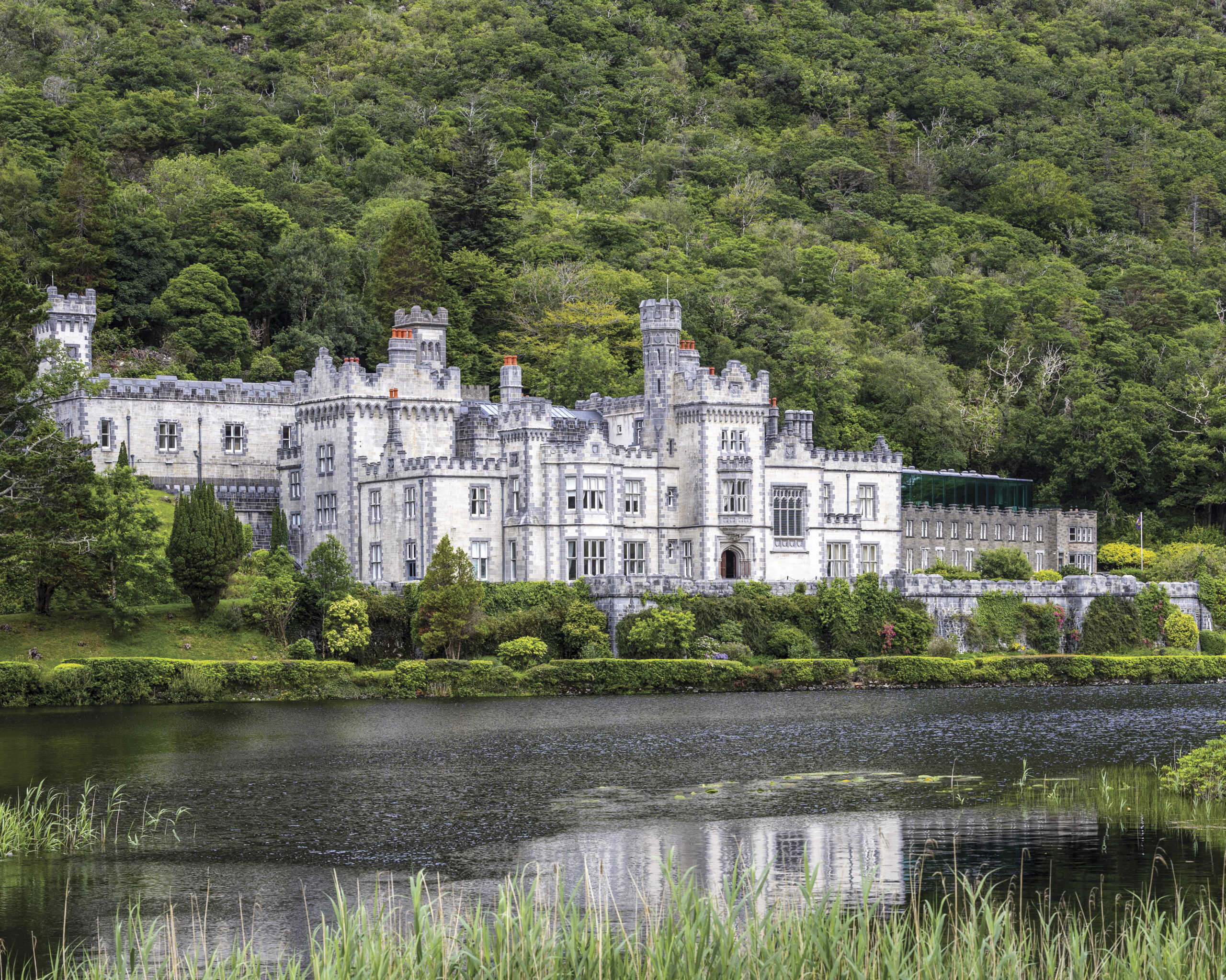 A stately gray stone castle with turrets and battlements sits along the edge of a calm lake, framed by manicured greenery and backed by a dense, verdant hillside.