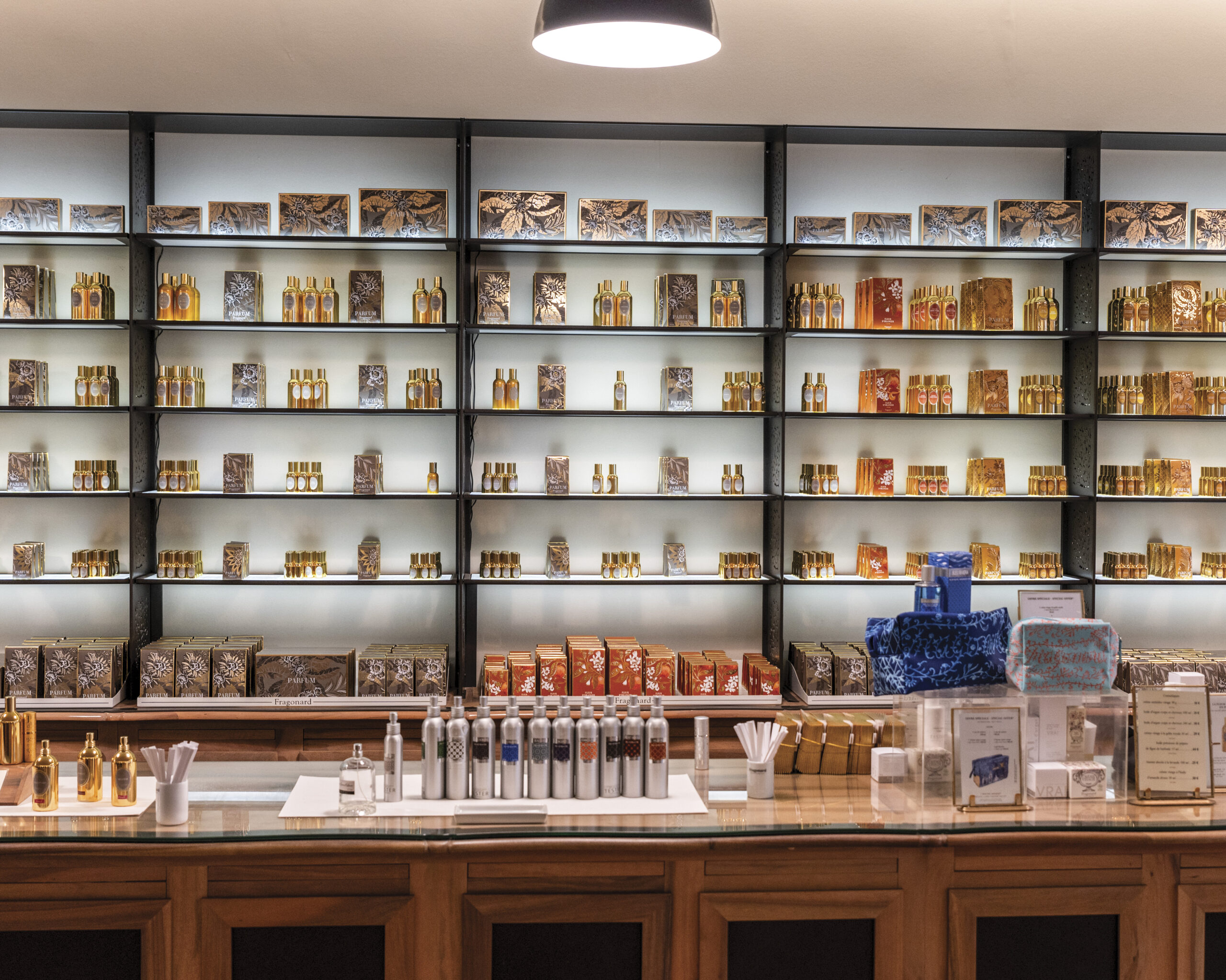 Elegant boutique interior with wall-to-wall shelving displaying rows of perfume bottles and boxed fragrances in warm amber tones, with a central counter arranged with testers, scent strips, and gift-ready packages.